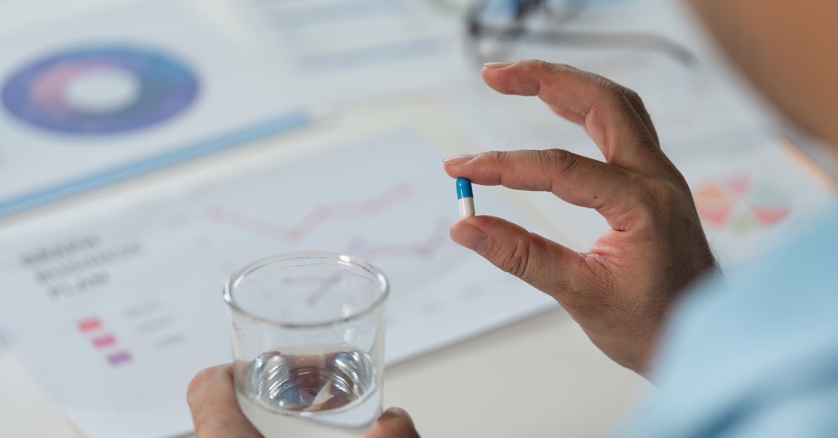 A person holds a blue and white pill in one hand and a glass of water in the other, with business charts in the background.