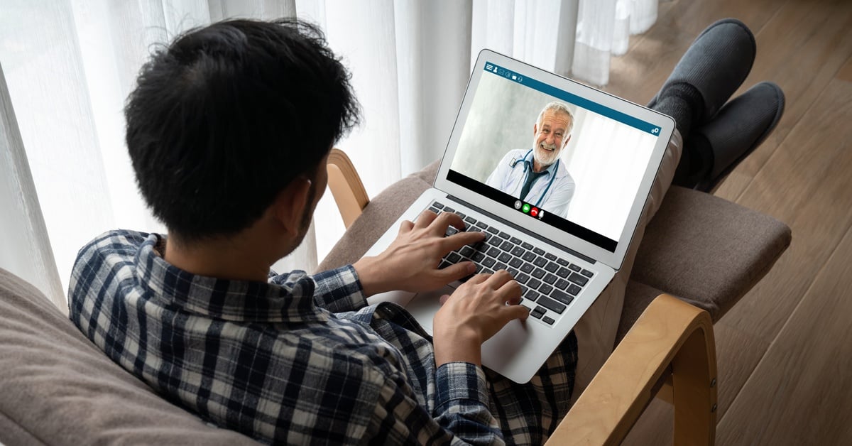 A man in a plaid shirt sits on a lounge chair, using a laptop for a telehealth video call with a smiling doctor.