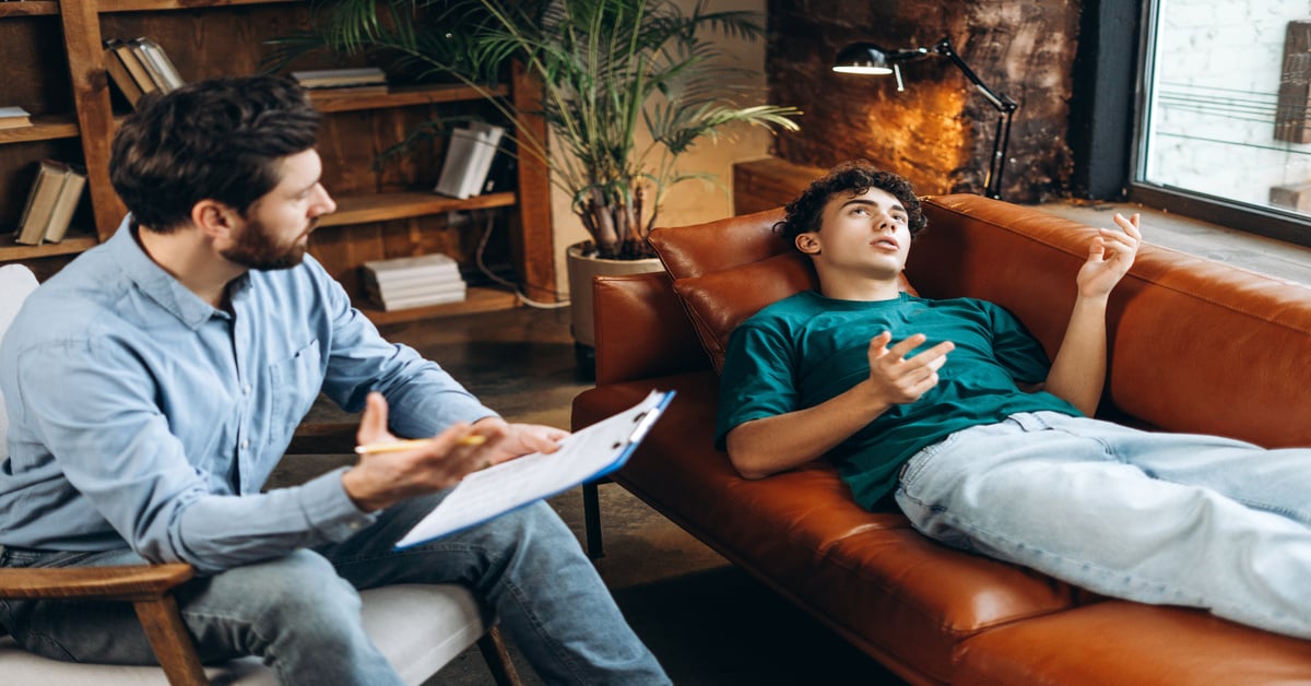 A young man lies on a brown couch during an anxiety therapy session, talking to a therapist who holds a clipboard.