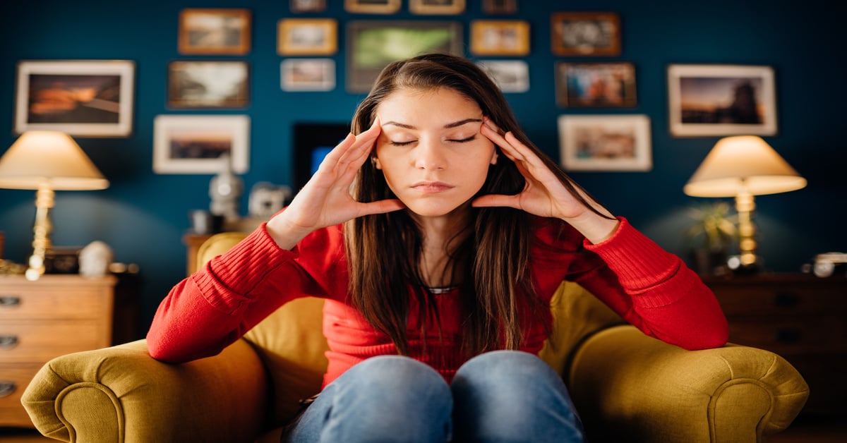 A young woman dealing with anxiety sits in a yellow chair with her hands on her head. Behind her is a blue wall with photos.