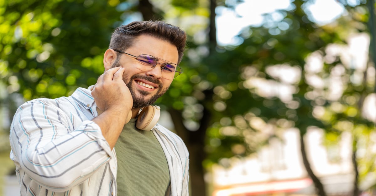 A man wearing glasses and headphones around his neck holds his hand to his face, a sign of jaw tension.