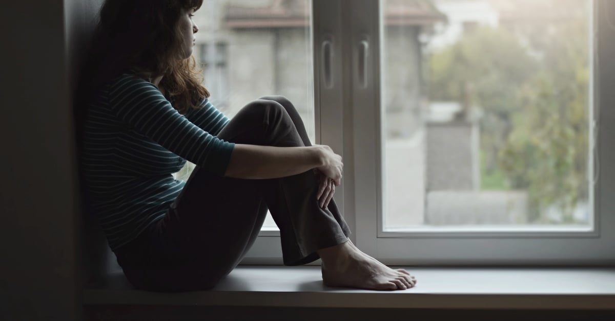 An isolated and depressed woman sits alone on a windowsill, hugging her knees while gazing out at neighboring houses.