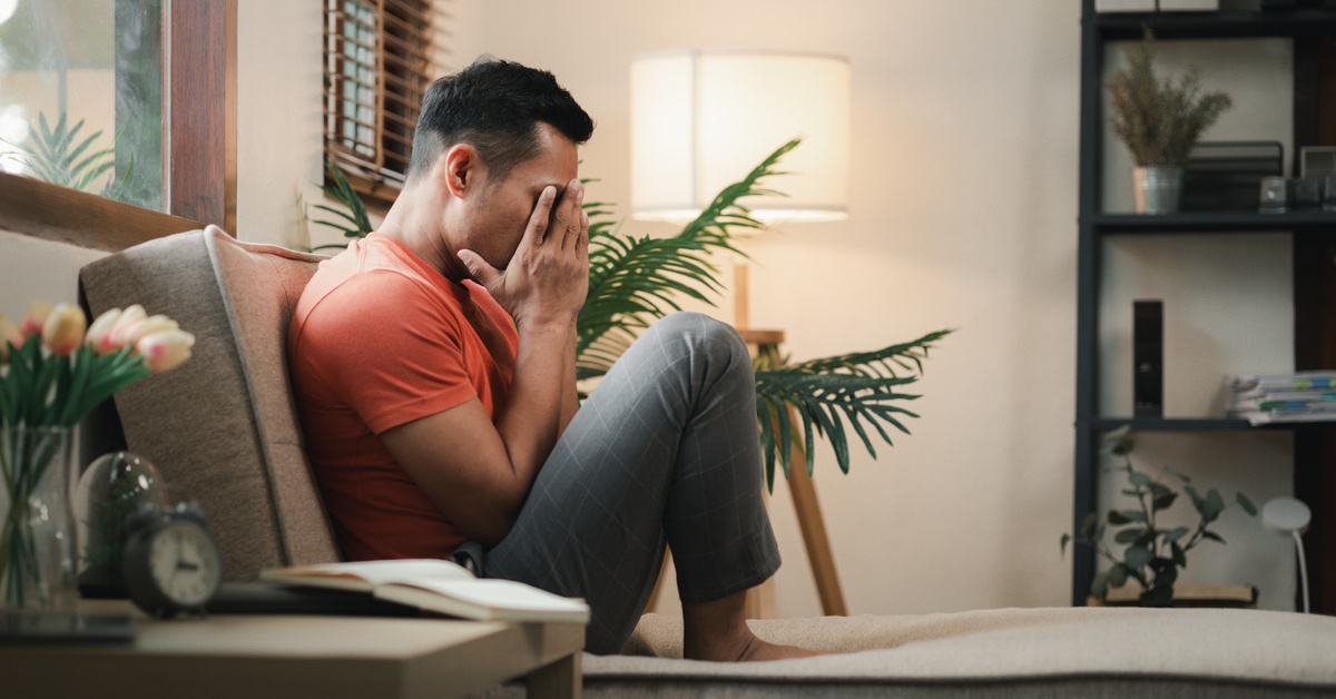 A stressed man in a red shirt sits on a couch, his hand covering his face, in a modern living room setting.