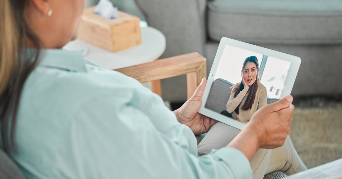 A therapist in a light blue shirt holds a tablet during a telehealth talk therapy session, speaking with a client on screen.