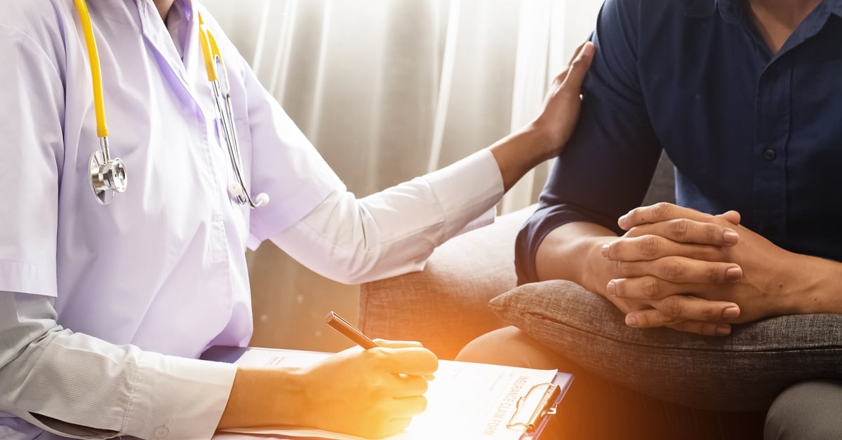 A psychiatrist in laboratory apparel places a comforting hand on their patient as they take notes on a clipboard.