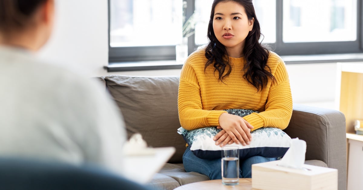 A woman in a yellow sweater sits on a couch with a pillow in her lap and talks to her therapist across from her.