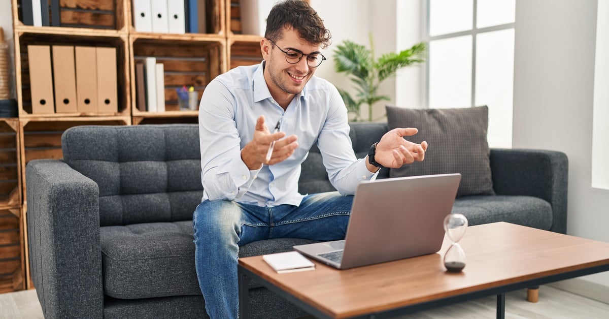 A smiling man sits on a sofa, gesturing as he speaks to his laptop during an online therapy session.