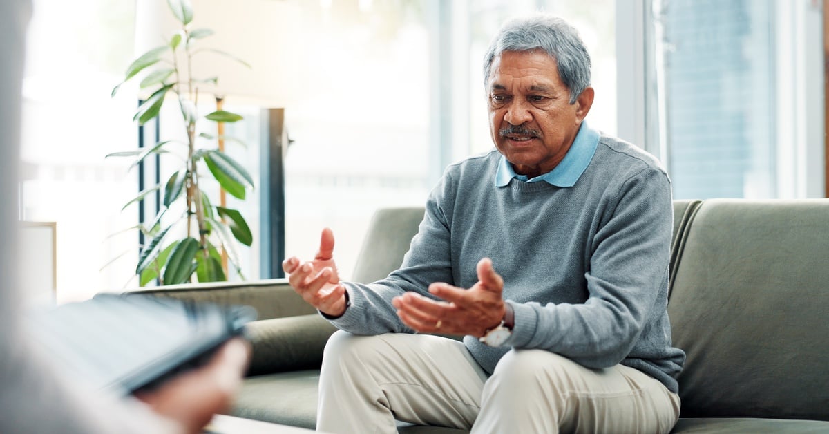 A senior man sits on a gray sofa as he talks with his hands. He wears a gray sweater with a blue collared shirt underneath.