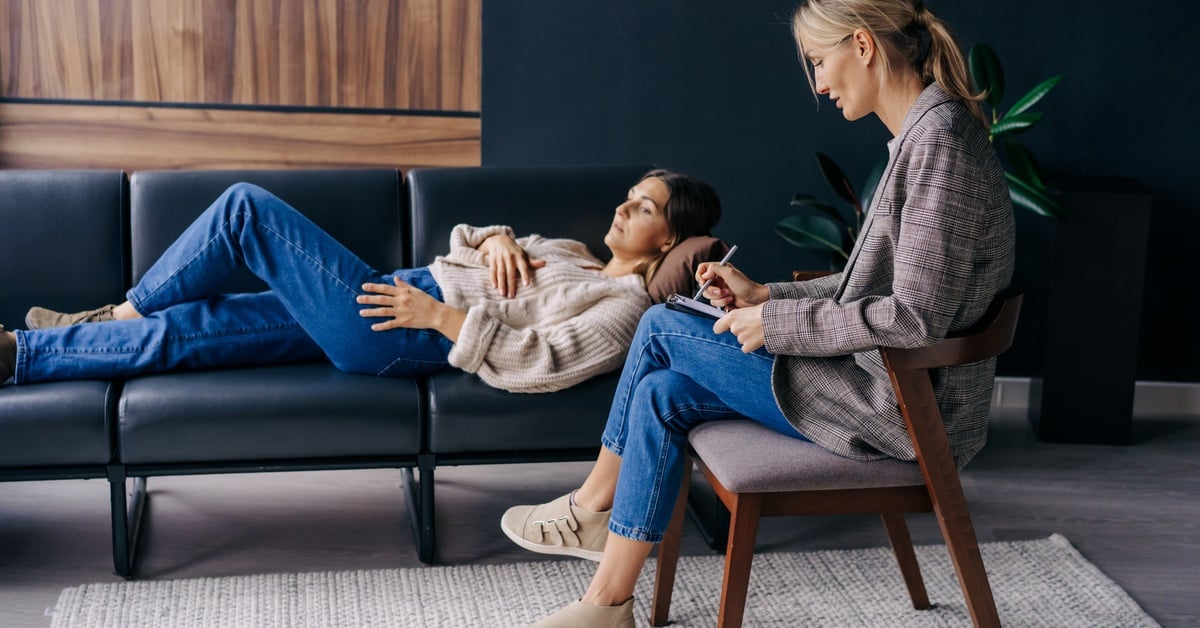 A woman in a modern office lies on her back, while another woman in professional attire sits nearby, taking notes.