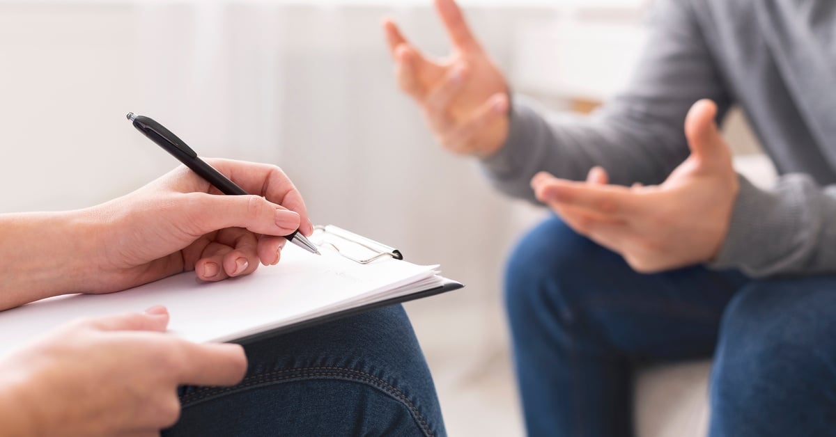 A close-up of two sets of hands. One person is taking notes on a clipboard, while the other gestures as they talk.
