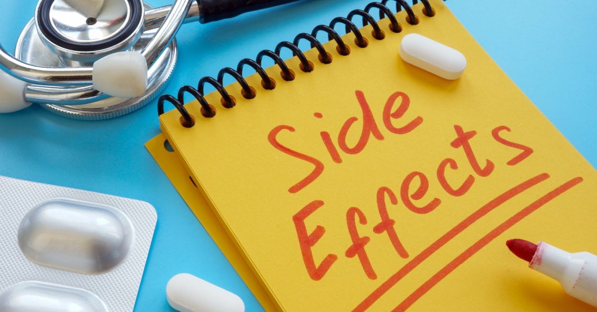 A yellow notepad with “side effects” written in red marker sits beside pills, a blister pack, and a stethoscope on a blue surface.