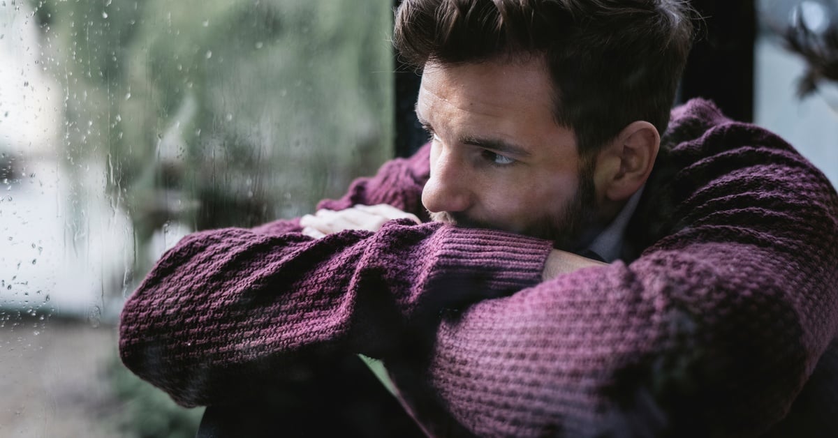 A young man, wrapped in a purple sweater, looks depressed and moody as he gazes out of a rainy window in winter.