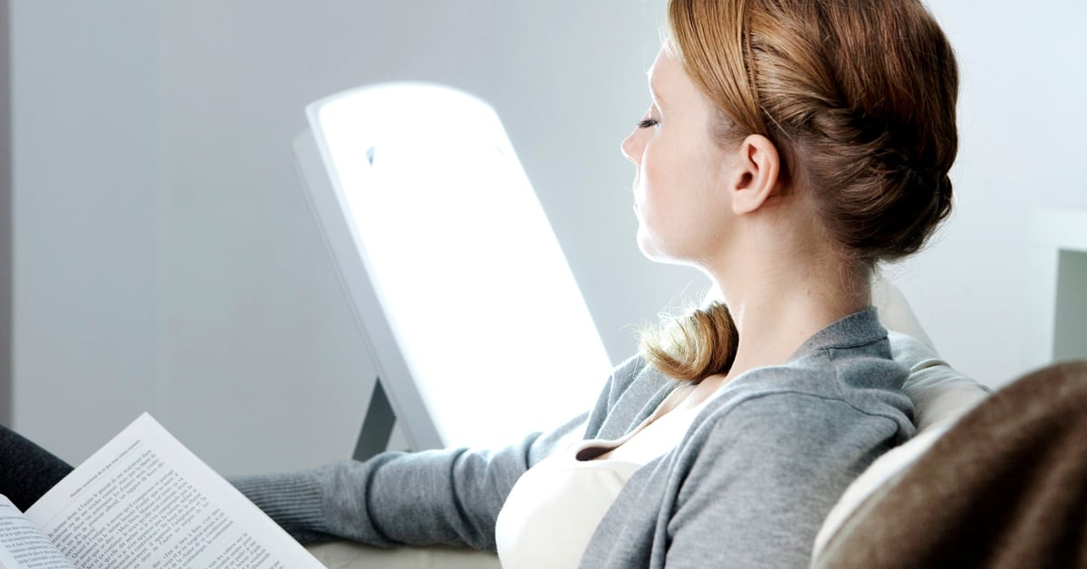 A woman tries to overcome seasonal depression by sitting near a bright therapeutic lamp while reading.