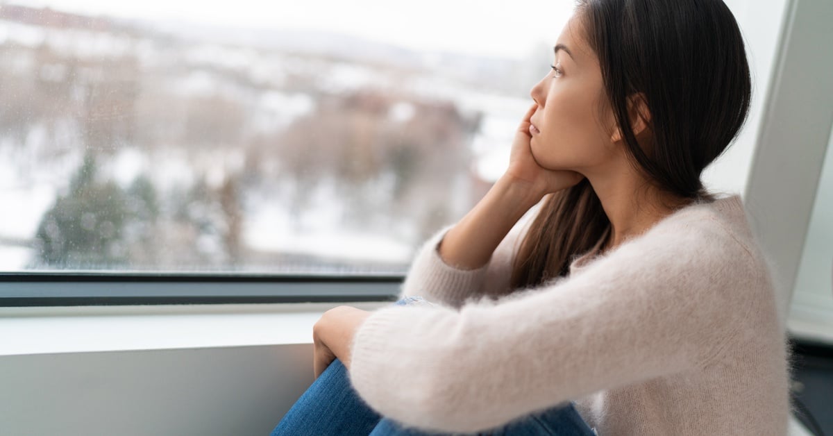 A young woman with seasonal affective disorder sits by a window on a gray winter day, her expression contemplative and melancholic.