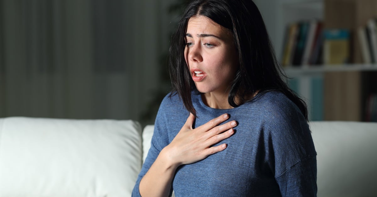 A woman with black hair and blue sweater touches her chest in distress with an anxious look on her face.