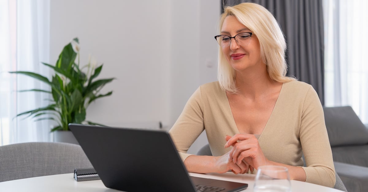 A middle-aged woman with blonde hair and glasses smiles as she engages in teletherapy on her laptop at home.