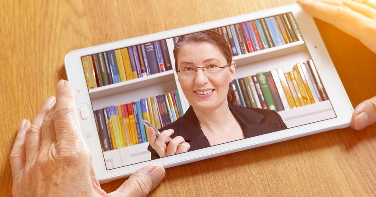 A pair of hands holds a tablet displaying a smiling therapist with glasses in front of a colorful bookshelf.
