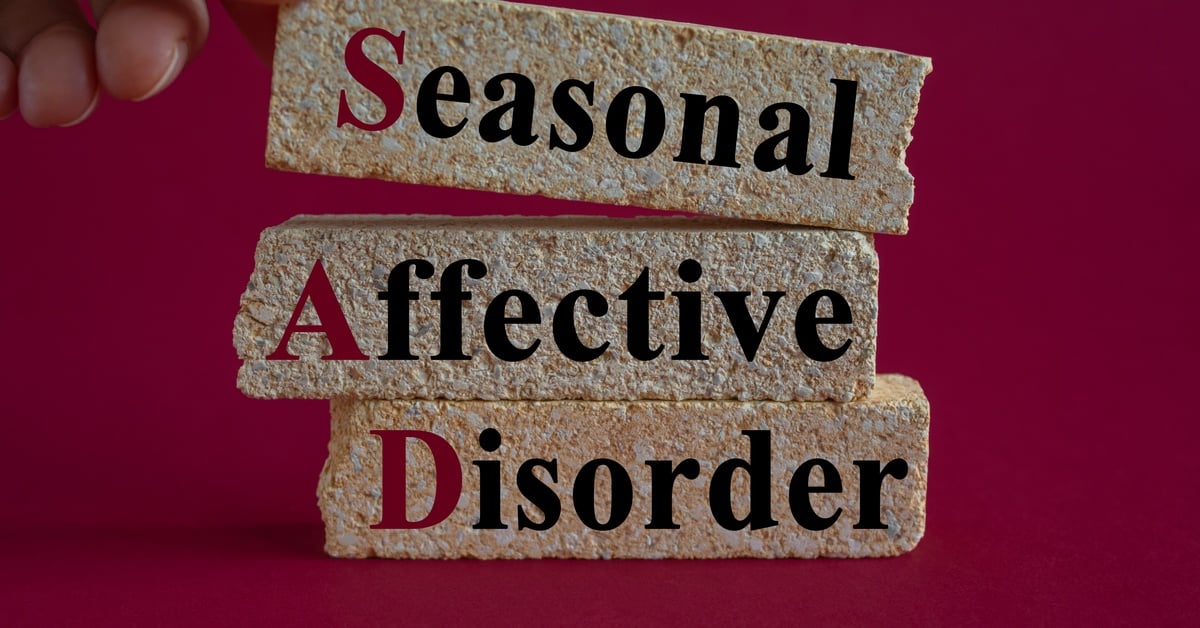 A person's finger touches stacked brick pieces spelling "Seasonal Affective Disorder" against a deep red background.