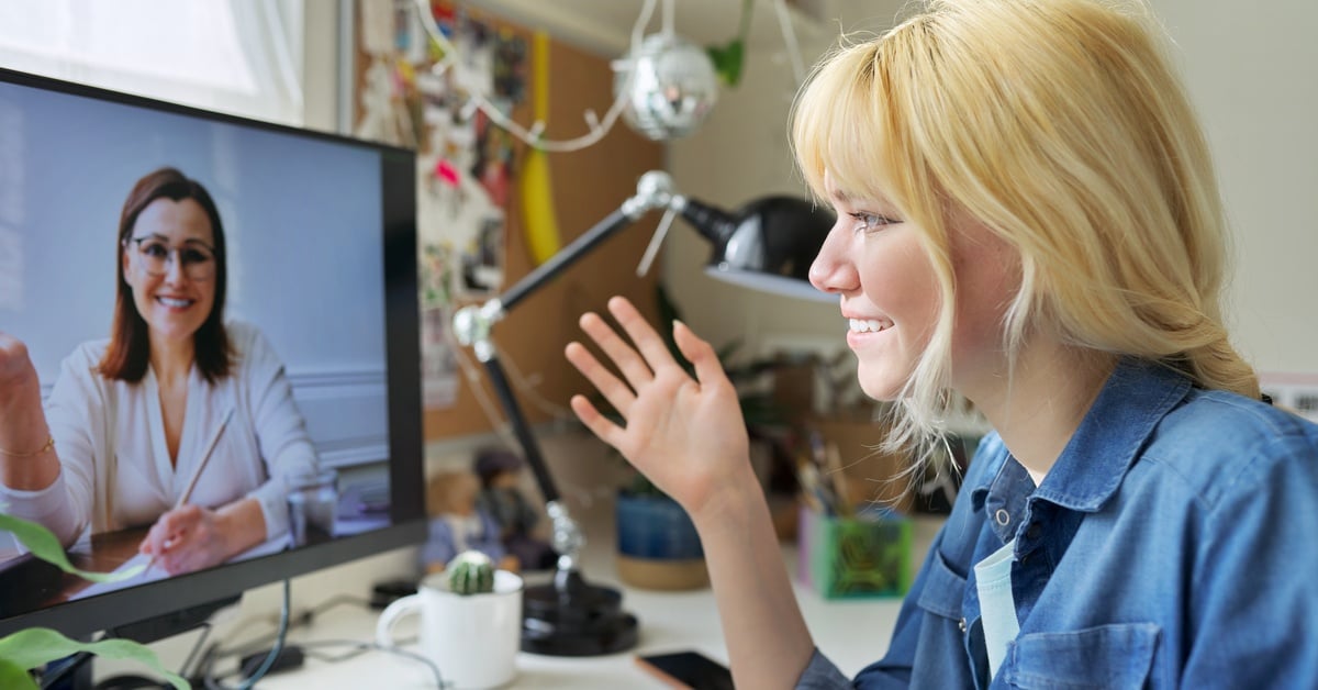 A young woman in a denim shirt smiles while speaking with a mental health professional during a telehealth psychiatry session.