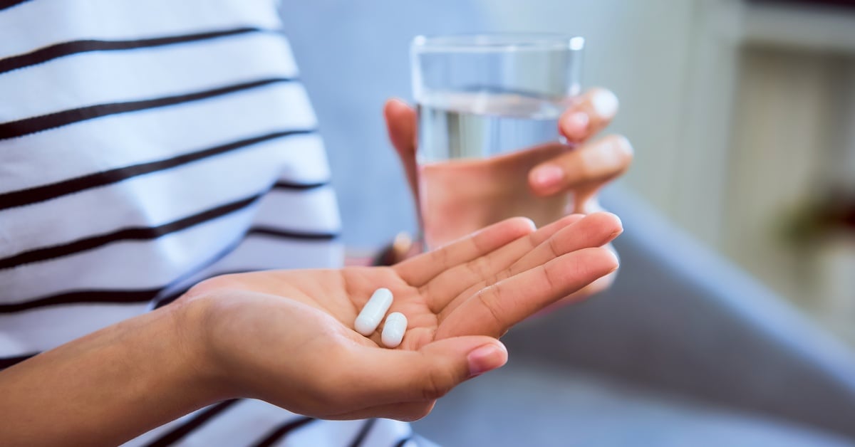A close-up of a person's hand holding two pills. They also hold a glass of water and wear a black and white striped shirt.