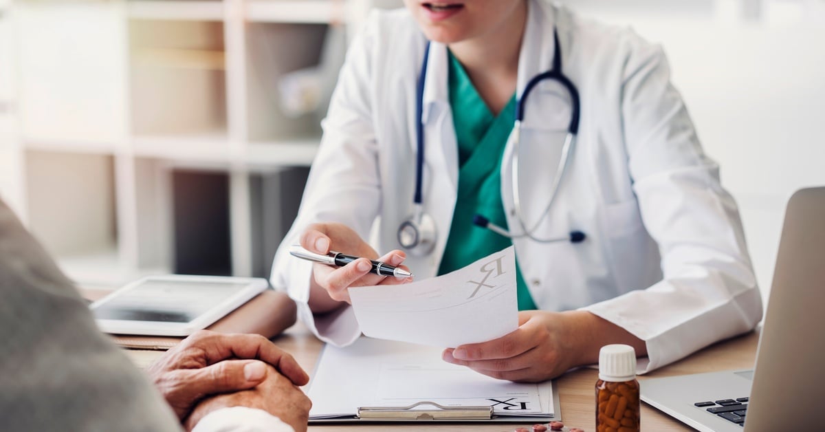 A patient sits across from a doctor as the doctor prescribes psychiatric medication to the patient on a script pad.