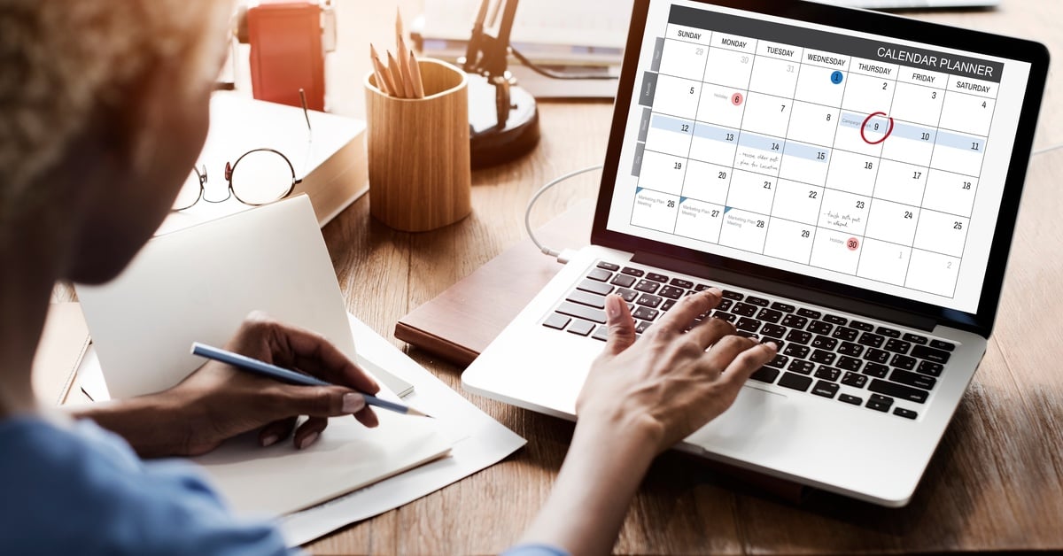 A person views a digital calendar planner on a laptop screen while taking notes at a wooden desk with office supplies.