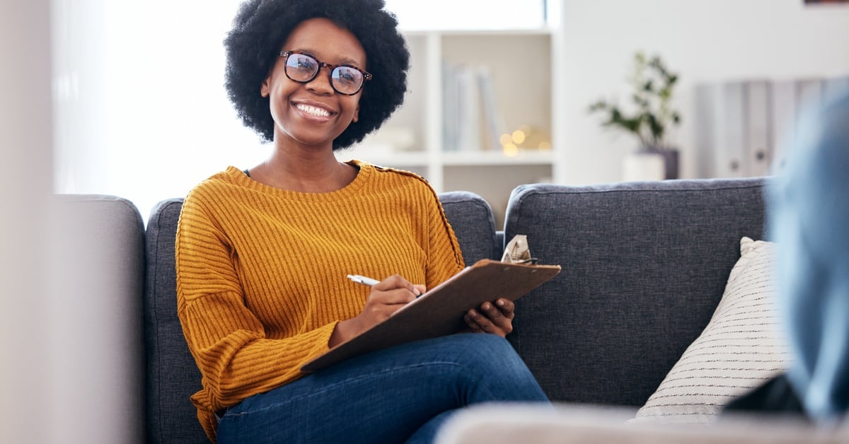 A Black woman therapist wearing a burnt orange sweater smiles at a patient while taking notes during a therapy session.