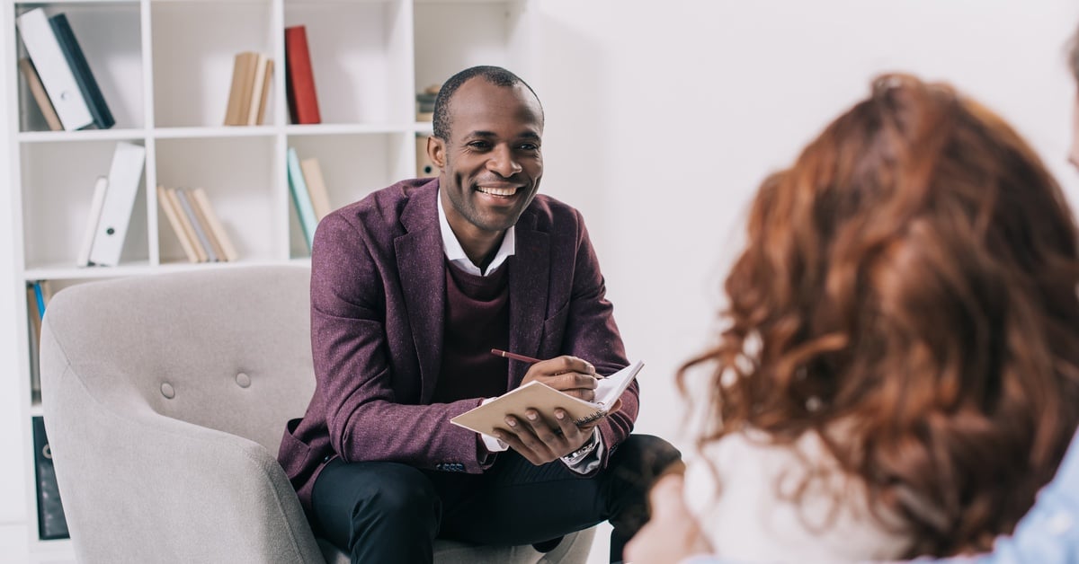 A Black male psychiatrist smiles while talking to his patients in an office. He writes down his assessments in a notepad.