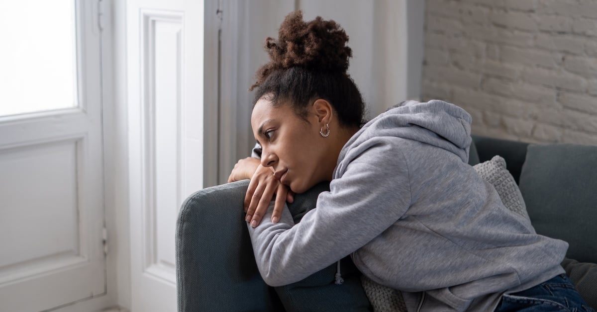A woman in a gray hoodie sits on a couch by a bright window, leaning on the armrest and staring outside with a reflective expression.