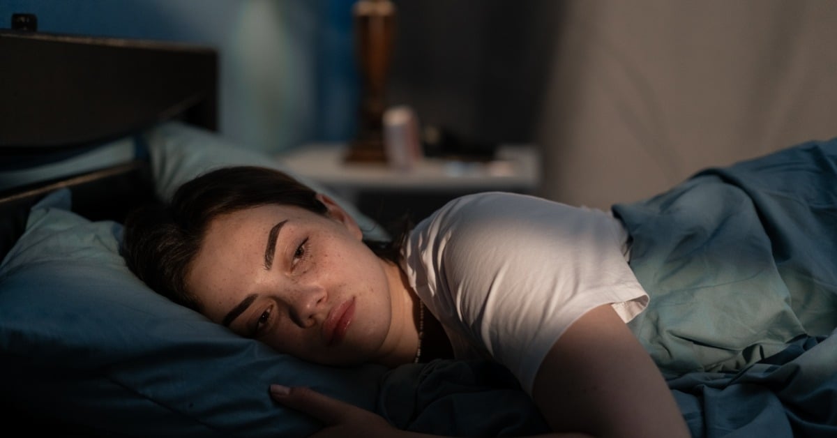 A woman lies in bed at night, looking tired and withdrawn, illuminated by soft lighting in a dim bedroom.