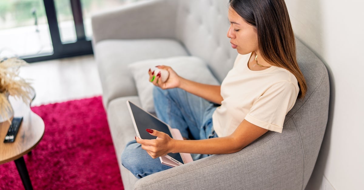 A woman sits on a couch at home holding her medication and a tablet while attending a virtual healthcare appointment.