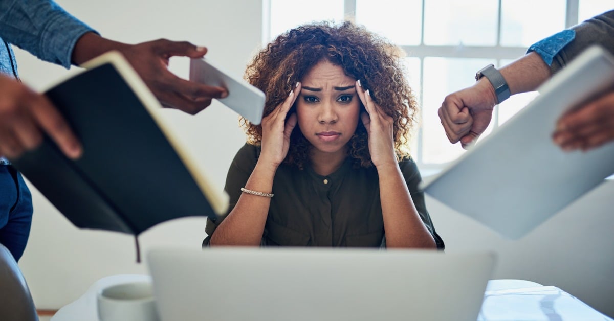 A young woman stares at a laptop and holds her temples as her coworkers hand her documents, illustrating professional burnout.