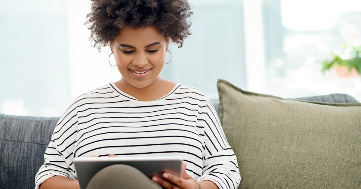 A young woman sits on a couch at home and smiles while using a tablet, attending a virtual therapy session.