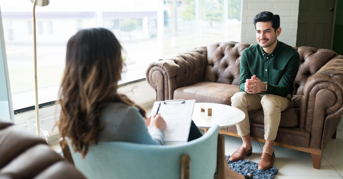 A therapist takes notes while speaking with a young adult man during an in-person counseling session in a bright office.