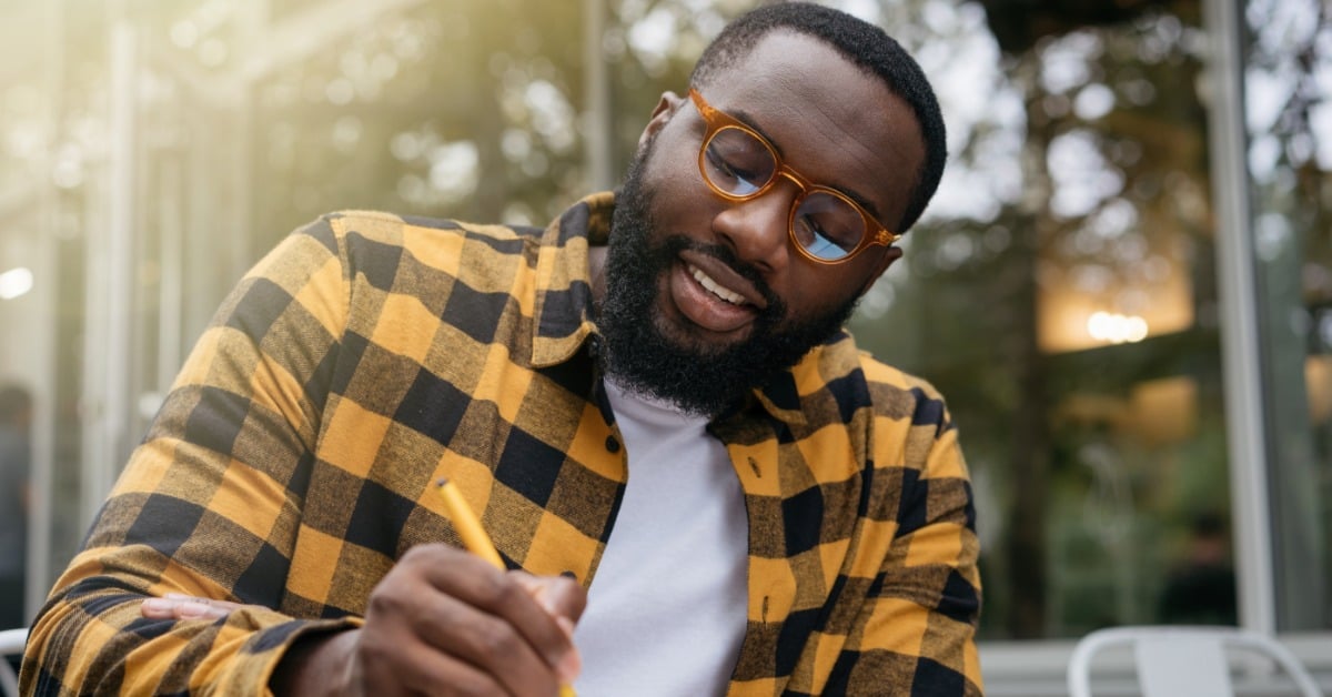 A young man wearing glasses and a plaid shirt takes notes before a therapy session while sitting outdoors with a notebook.