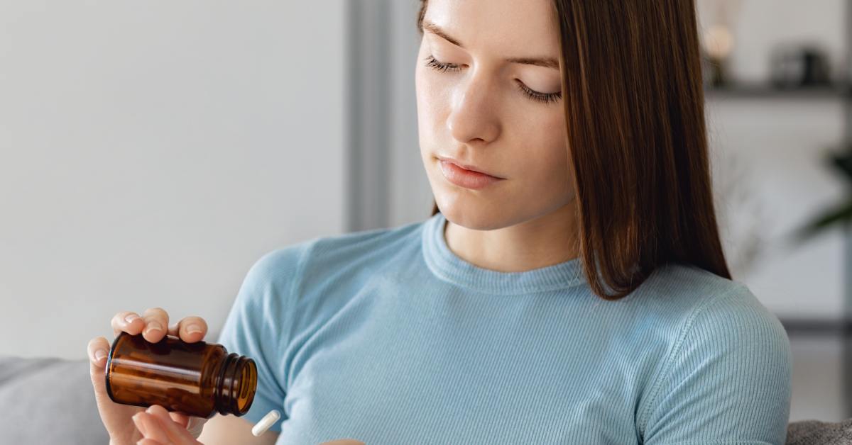 A young woman at home pours medication from a brown pill bottle into her hand while checking her antidepressant dose.