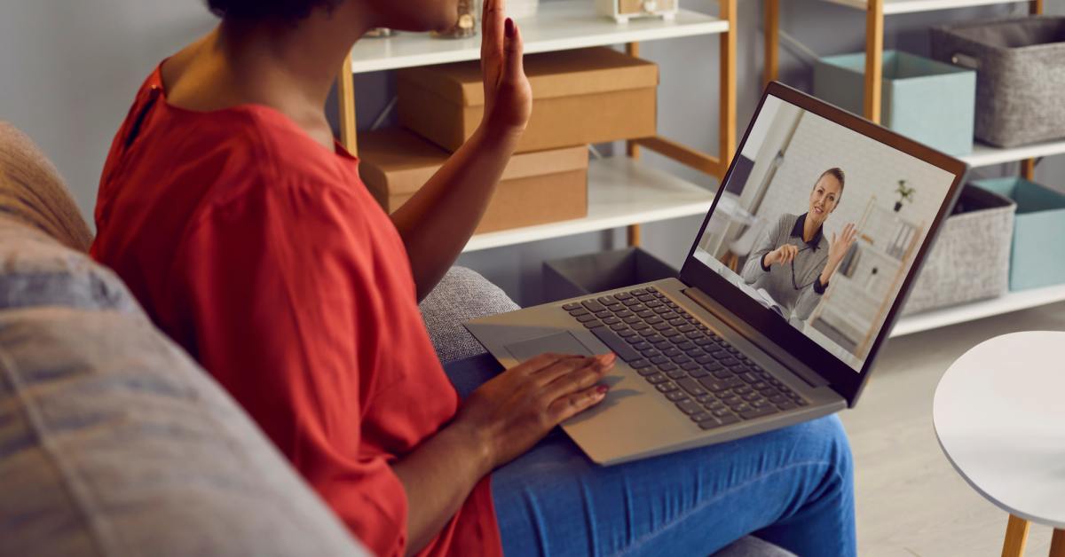 A woman sits on a couch and waves during a telehealth video call with a mental health provider on her laptop at home.