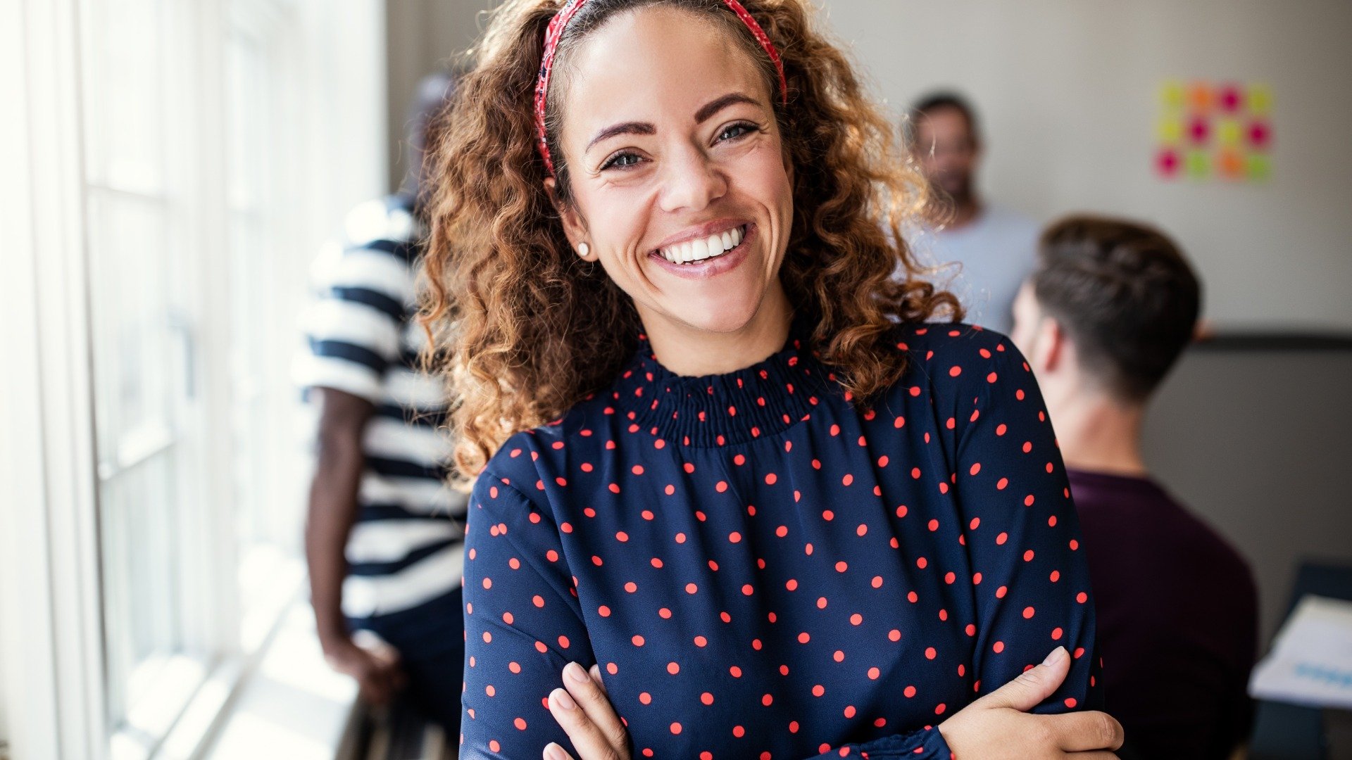 A smiling woman with curly hair, a red headband, and a polka-dot shirt stands with her arms crossed in a room with her peers.