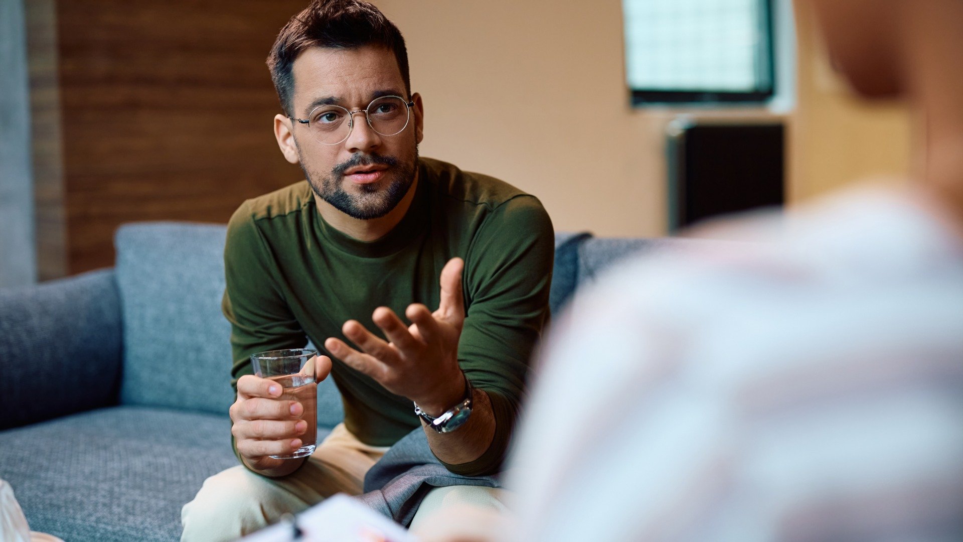 A man holds a glass of water and gestures while talking with a therapist during a mental health counseling session.