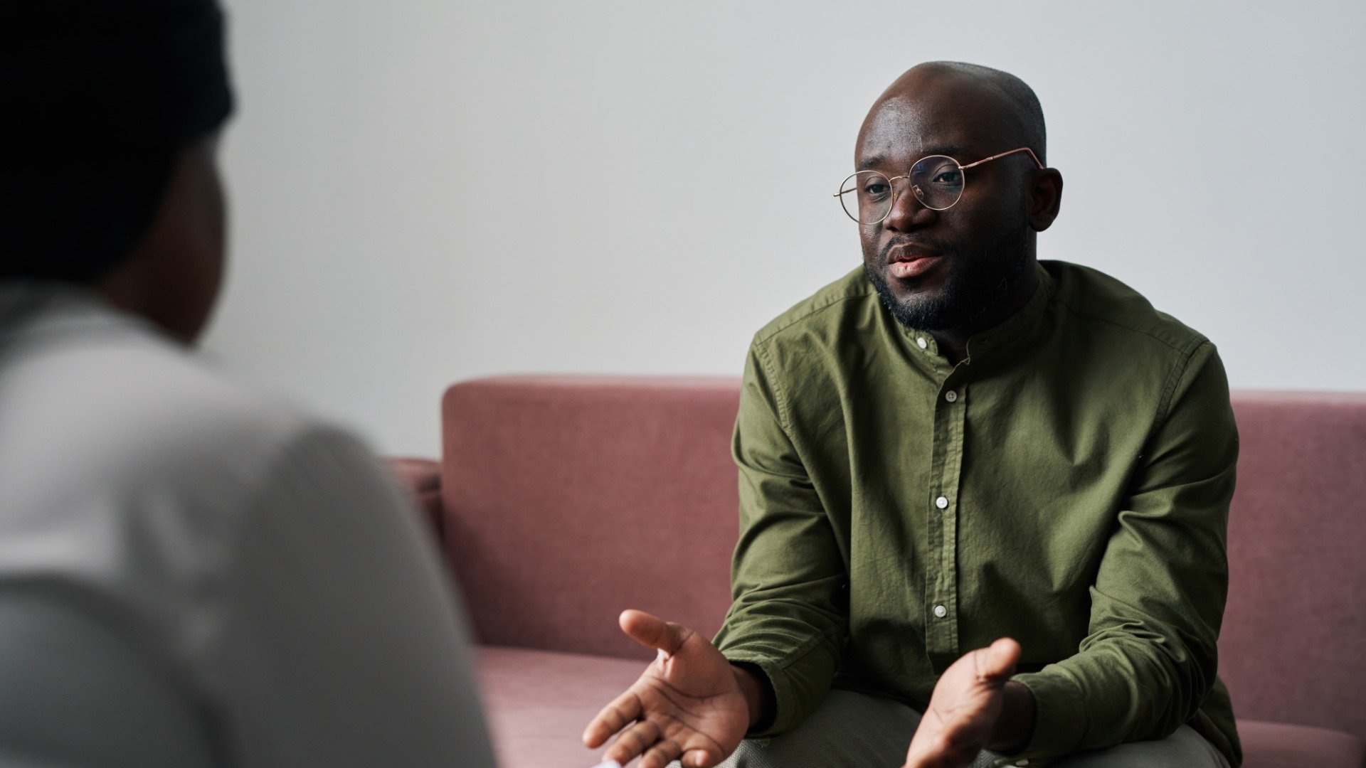 A man in a green shirt gestures while speaking with a therapist during a counseling session, seated on a couch in an office.