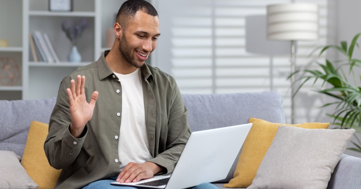 A man sits on a couch at home, smiling and waving during a virtual therapy session on a laptop in a well-lit living room.