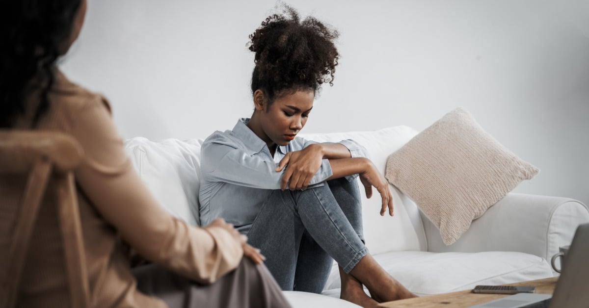 A young woman sits curled up on a couch during a therapy session, dealing with symptoms from anxiety and stress.