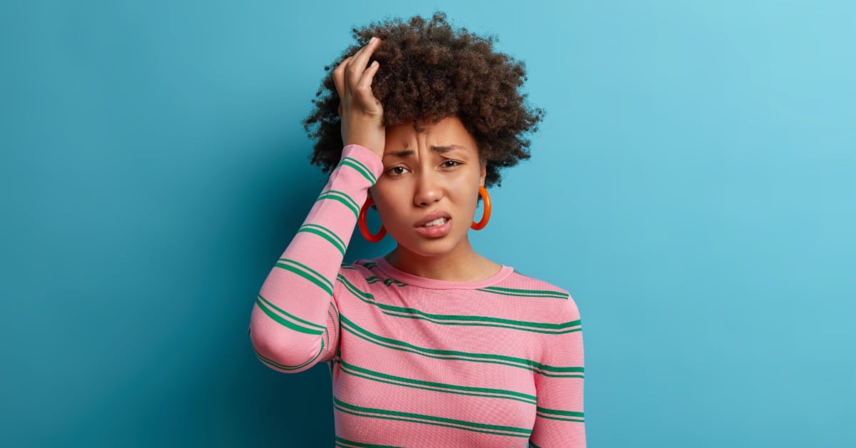 A young woman holds her head and looks overwhelmed against a blue background, expressing worry, confusion, or mental stress.