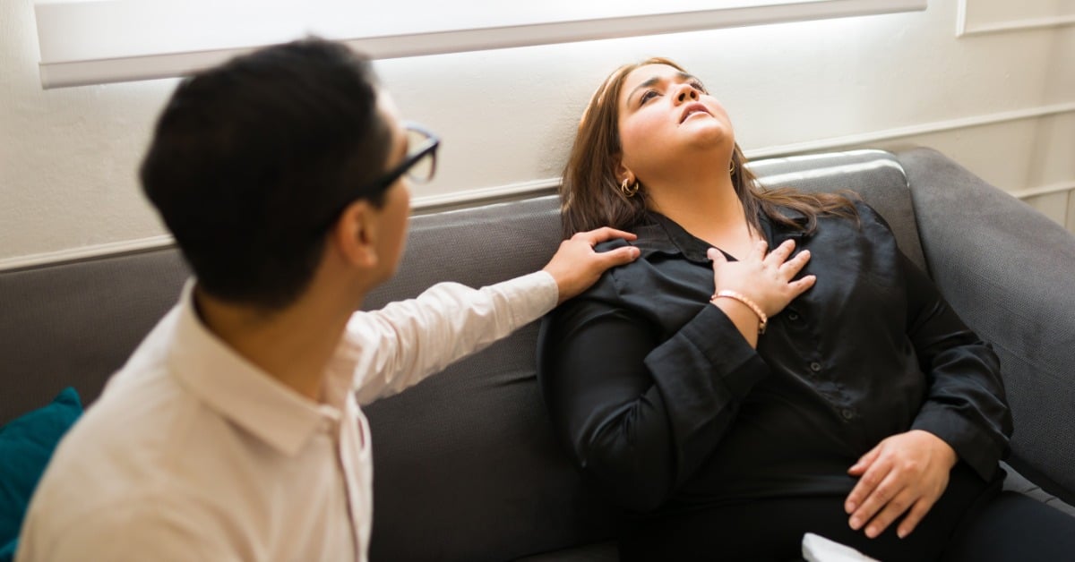 A distressed woman reclines on a gray couch holding her chest while a man in glasses reaches out to comfort her.
