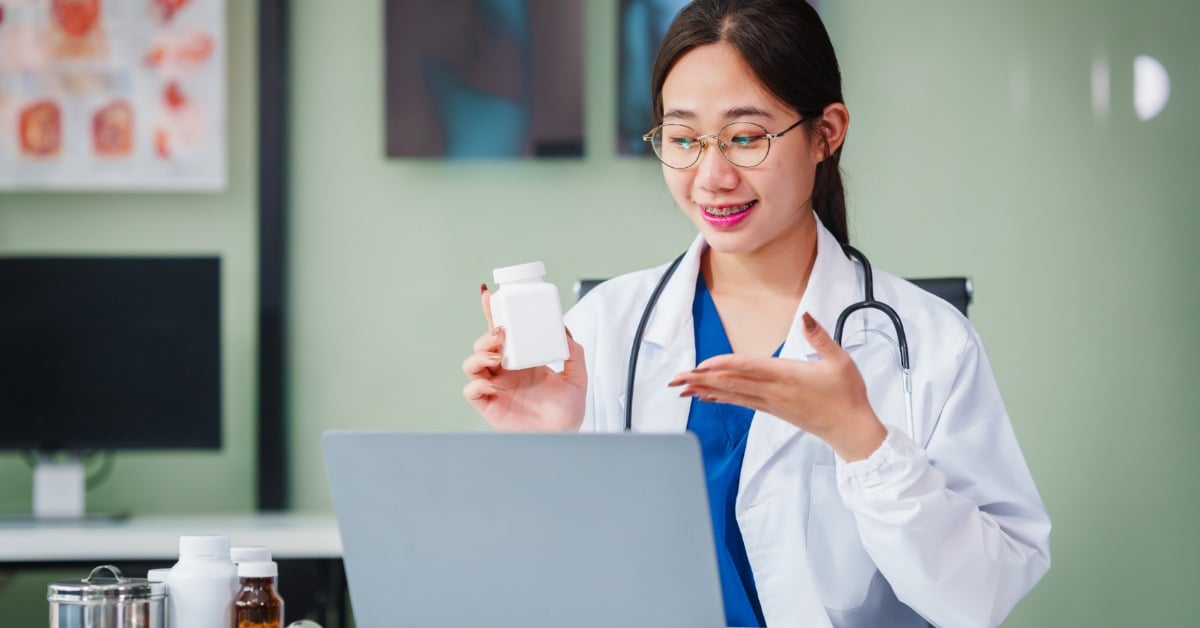 A smiling female doctor in a white coat holds a pill bottle during a telehealth video appointment on a laptop.