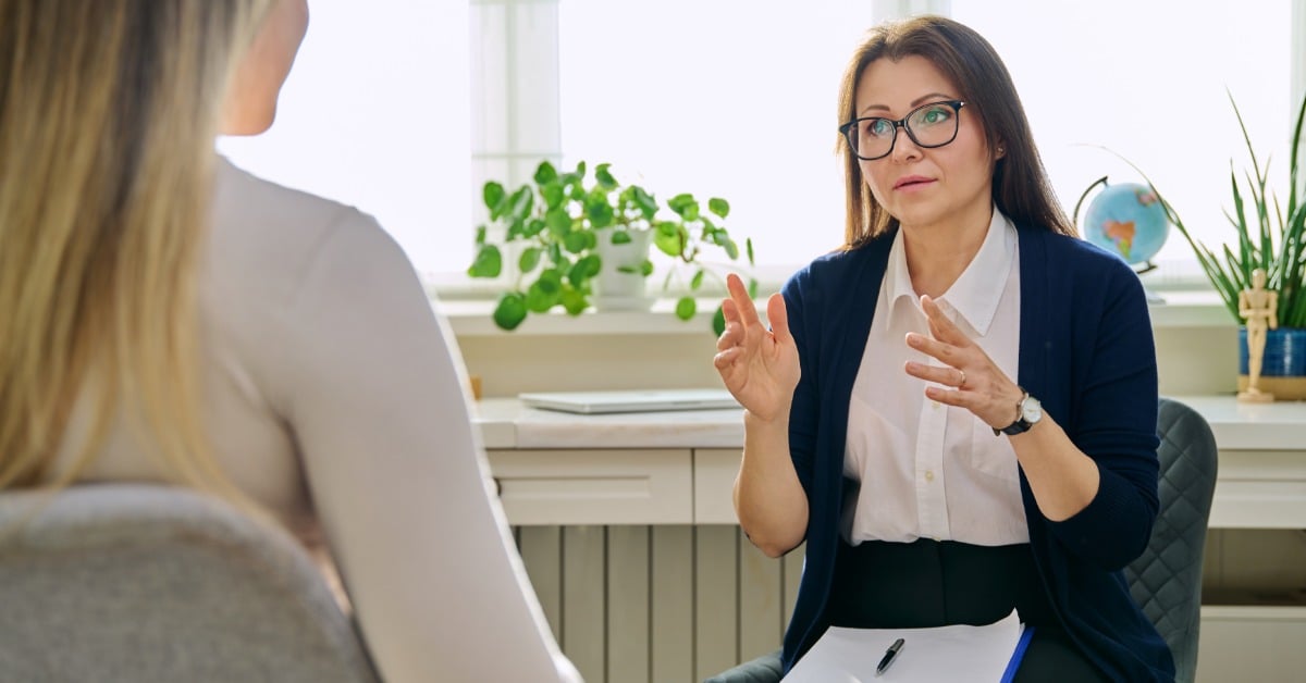 A psychiatrist speaks with a patient during a therapy session, taking notes while explaining something in a professional setting.