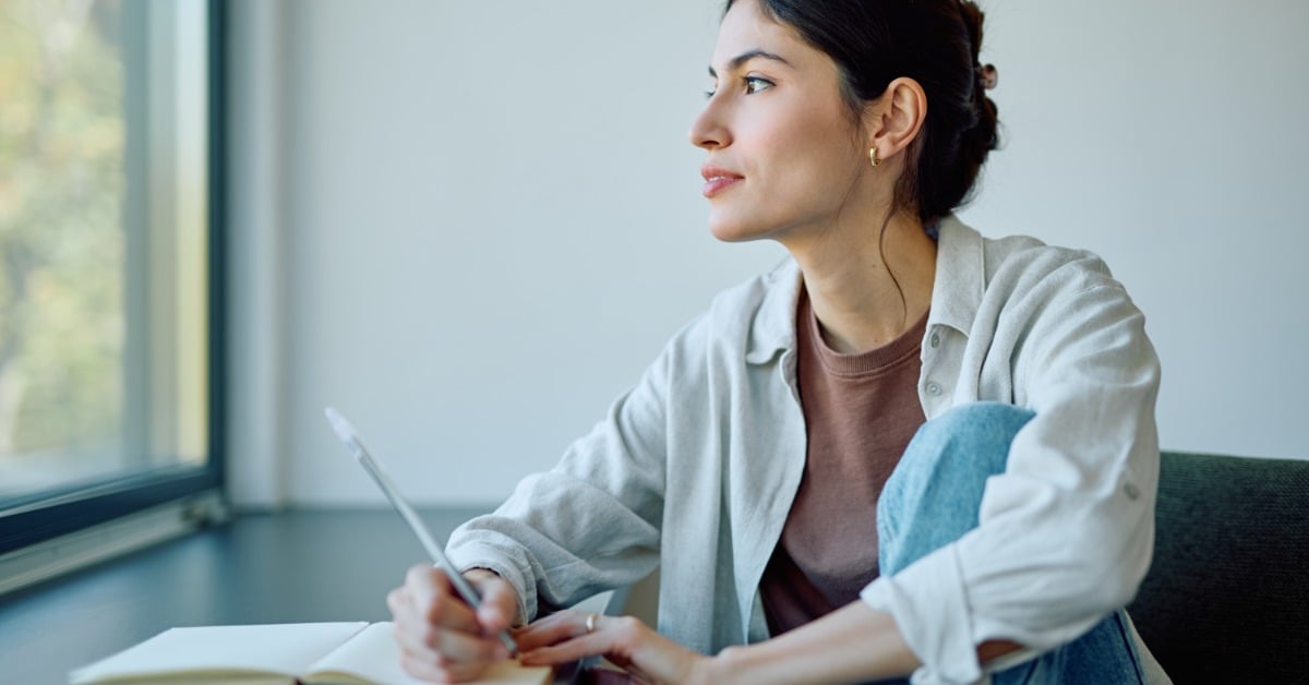 A woman sits by a window, writing in a notebook as she prepares notes and thoughts ahead of a psychiatry session.