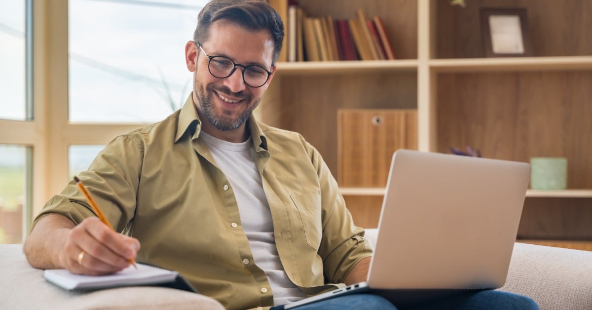 A smiling man with glasses takes notes on a notepad during a telehealth therapy session on his laptop at home.
