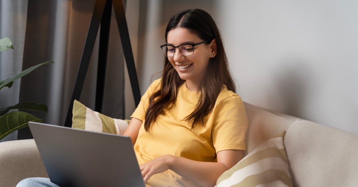 A smiling young woman with glasses in a yellow shirt on her couch at home, attending a telehealth therapy session on her laptop.