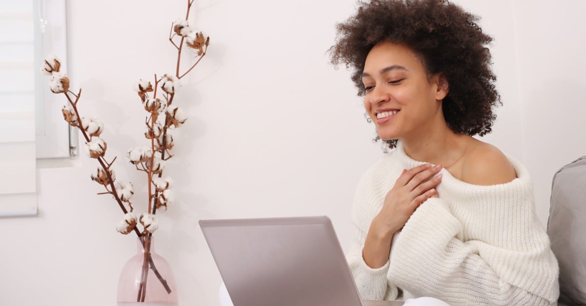 A smiling young woman sits comfortably on her couch with a laptop during a telehealth mental health session at home.
